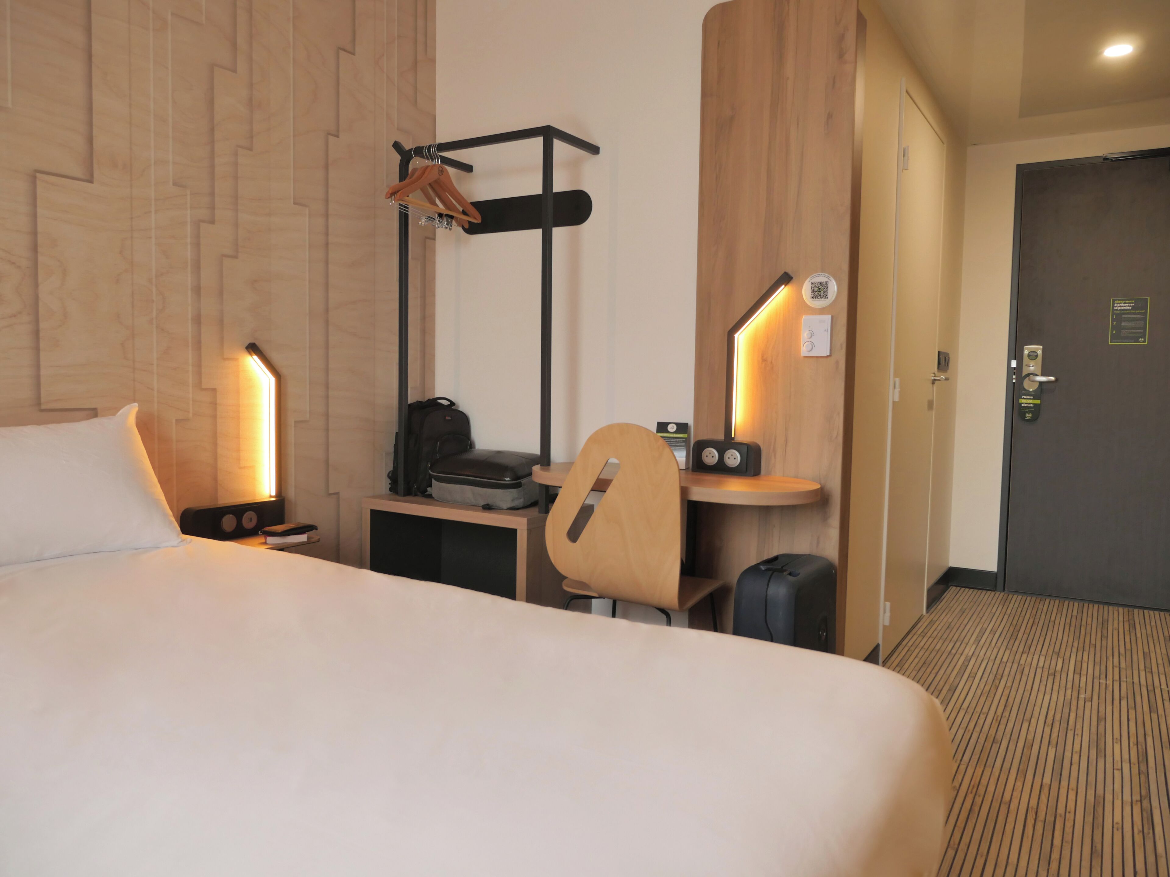 Habitación de hotel con cama, escritorio con silla, perchero, maleta y puerta con cerradura.
