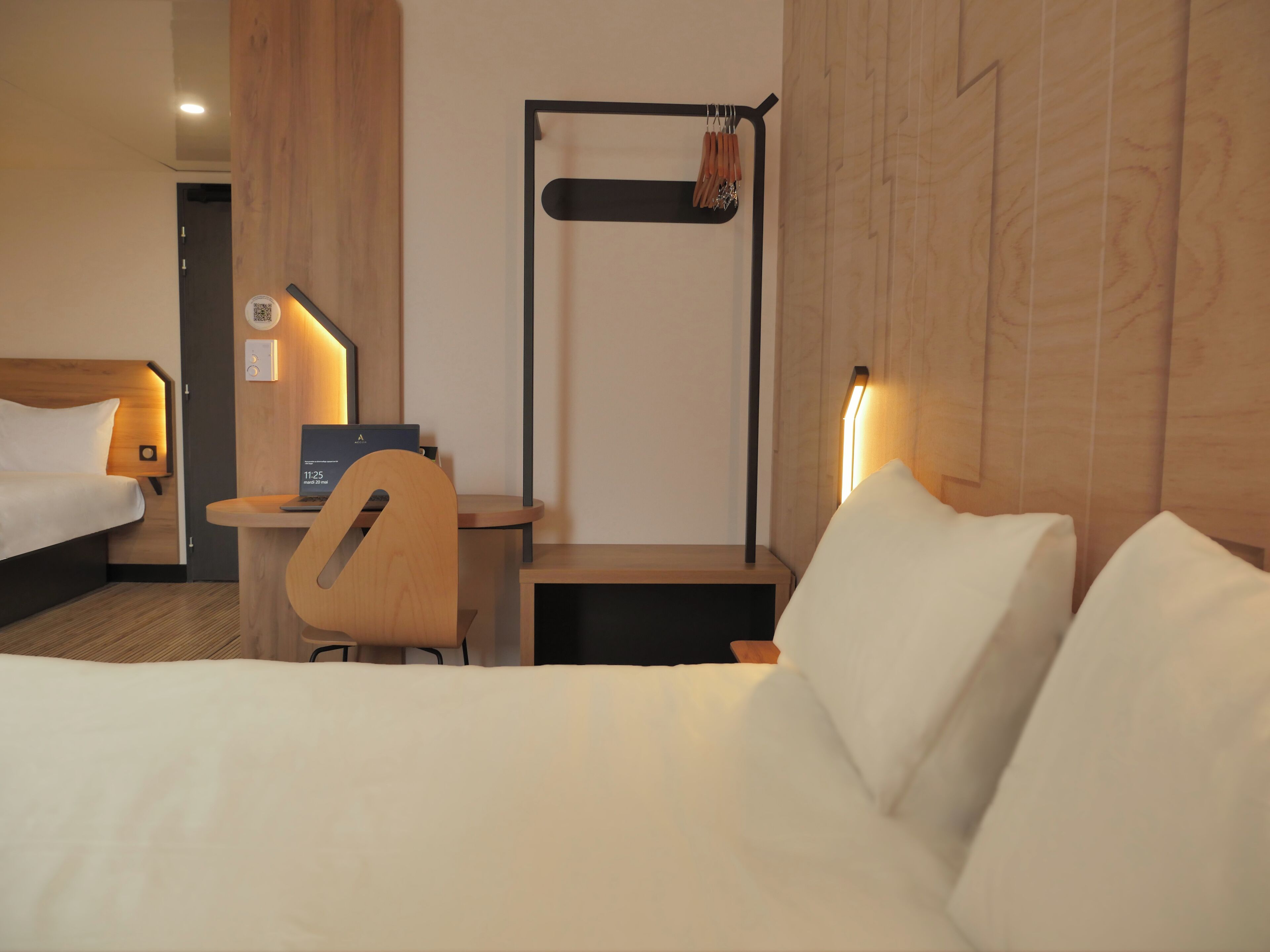 A hotel room with a bed, a desk, a chair, a small table, and a wall-mounted rack with clothes.
