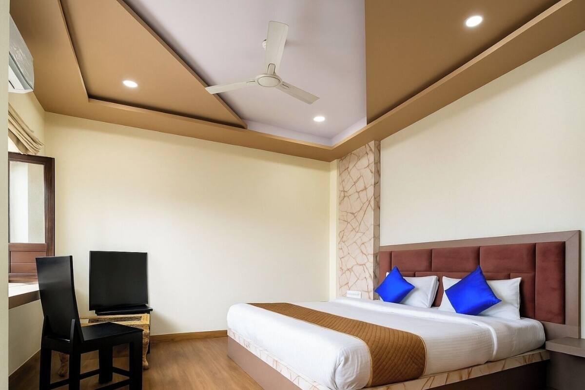 A hotel room with a bed, a desk with a chair, a ceiling fan, and a wall-mounted TV.