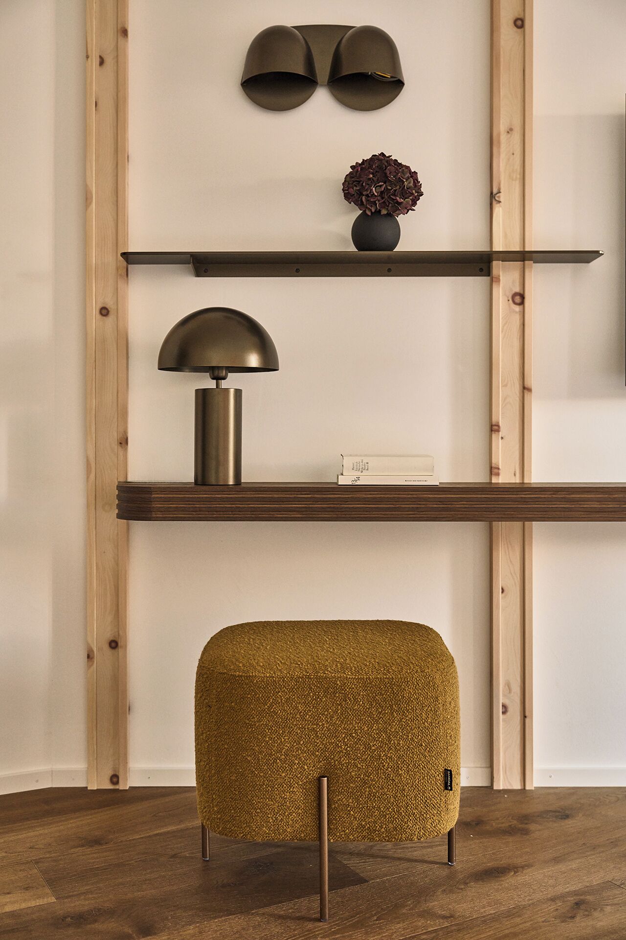 Una habitación con piso de madera, una repisa con lámpara, un florero con flores y un otomano texturizado.
