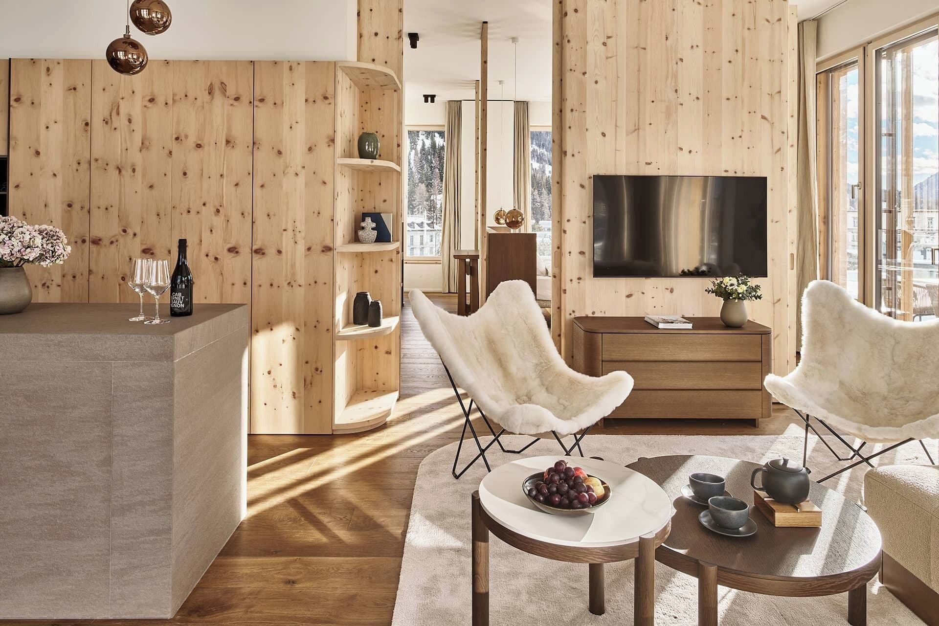 Un salon moderne avec des murs recouverts de panneaux en bois, un grand téléviseur à écran plat, deux fauteuils blancs, une table basse ronde avec des fruits et des tasses, et une petite table d’appoint avec une théière.