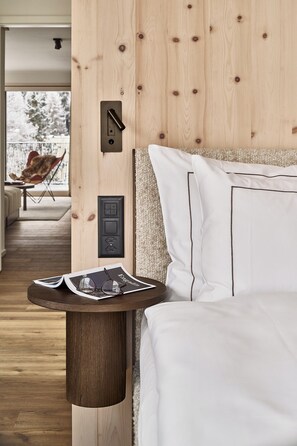 A bedroom with a wooden headboard, a bedside table with glasses and a magazine, and a view of a balcony with a chair and a snowy landscape.