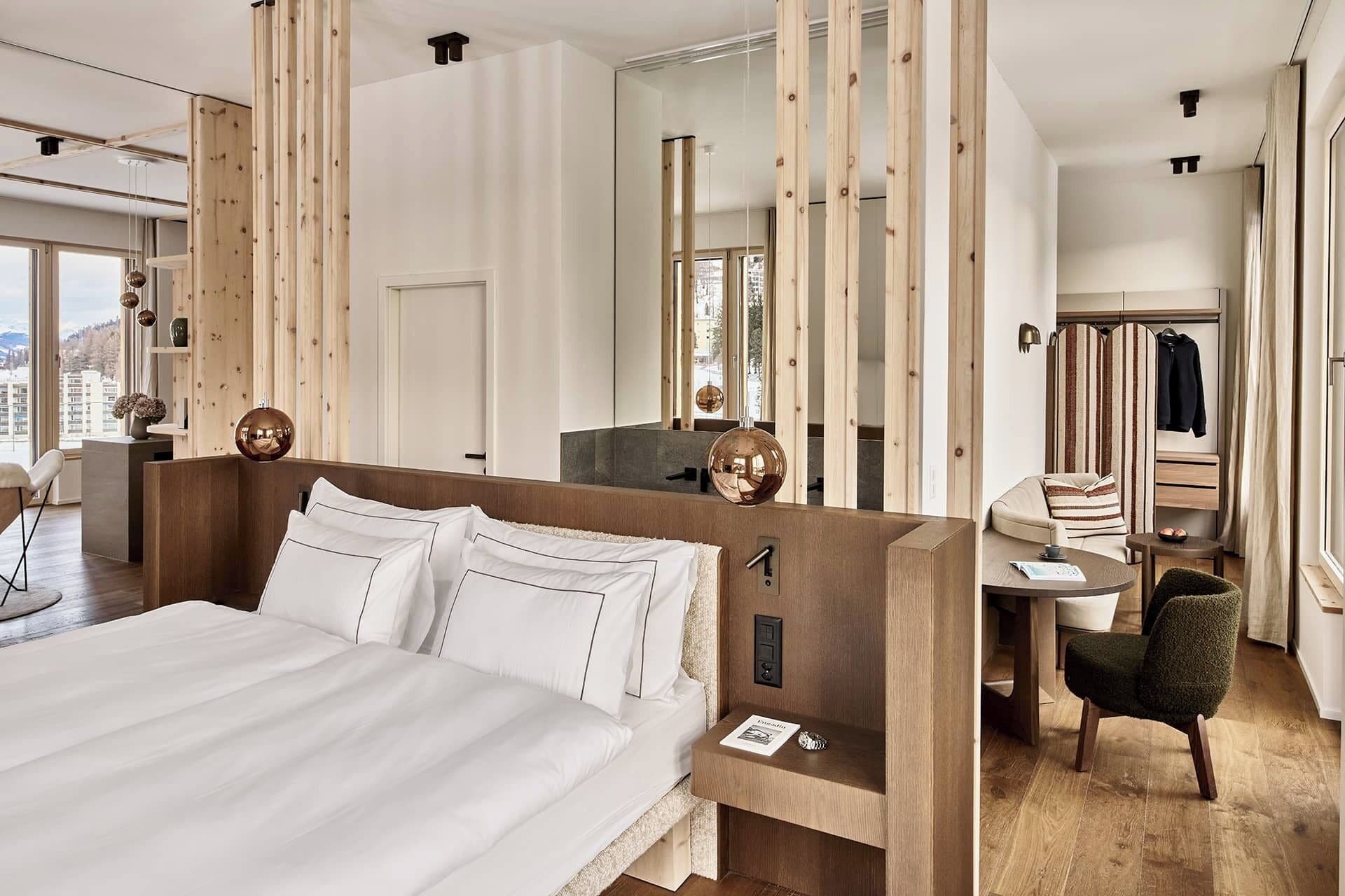 Quarto de hotel moderno com uma cama grande, uma escrivaninha e uma cadeira. Há um espelho na parede e uma janela com vista para a cidade.
