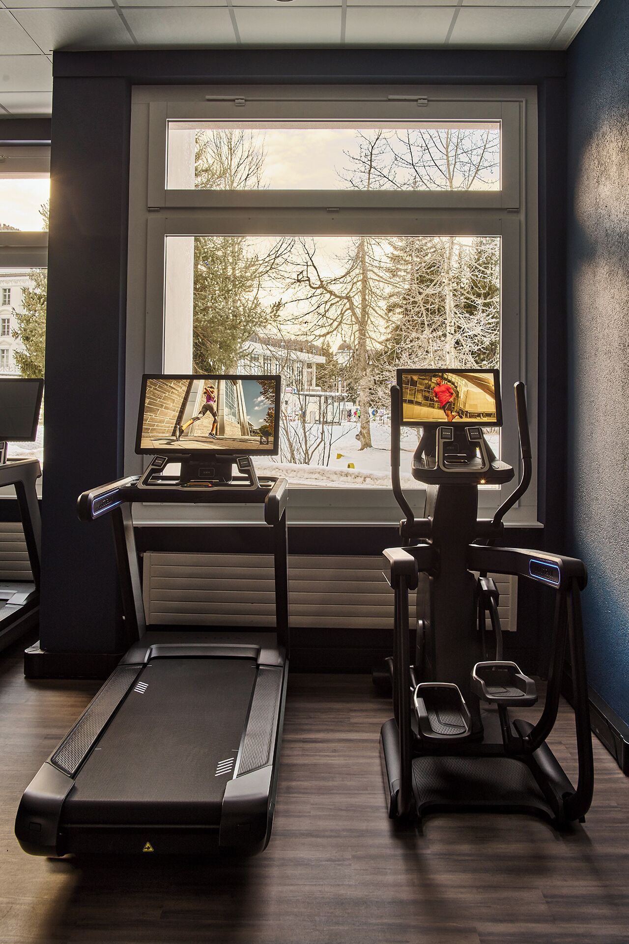 Ein Fitnessstudio mit Laufbändern, Crosstrainern und Ergometer, mit großen Fenstern, die einen Blick auf eine verschneite Landschaft bieten.