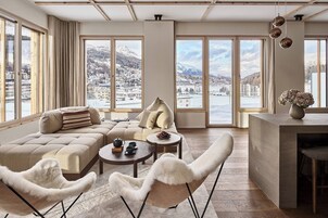 Un salón amplio con un gran ventanal que ofrece vistas a un paisaje nevado y edificios. Se observa un sofá, dos sillones, una mesa de centro y una mesita auxiliar con un jarrón de flores.
