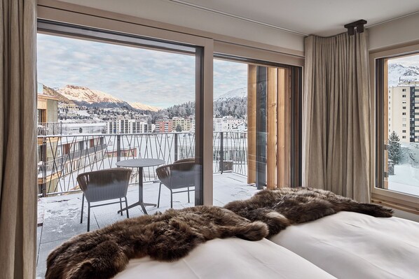 A hotel room with a large window offering a view of a snowy mountain and a cityscape. - The View (St. Moritz)