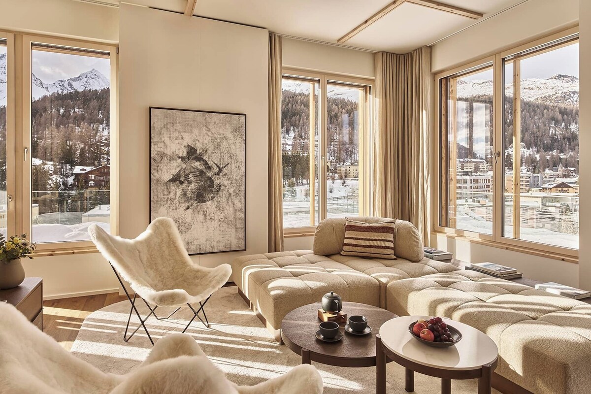 A modern living room with large windows, a plush sofa, a white chair, and a round table with a teapot and cups.