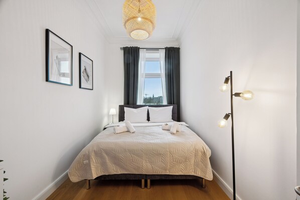 Una cama bien hecha con una colcha blanca, una lámpara de noche y una ventana con cortinas.