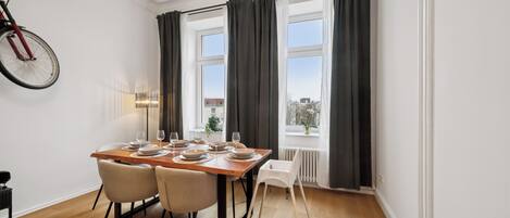 Un coin repas avec une table en bois dressée pour quatre personnes, deux chaises, un vélo et une fenêtre avec des rideaux.