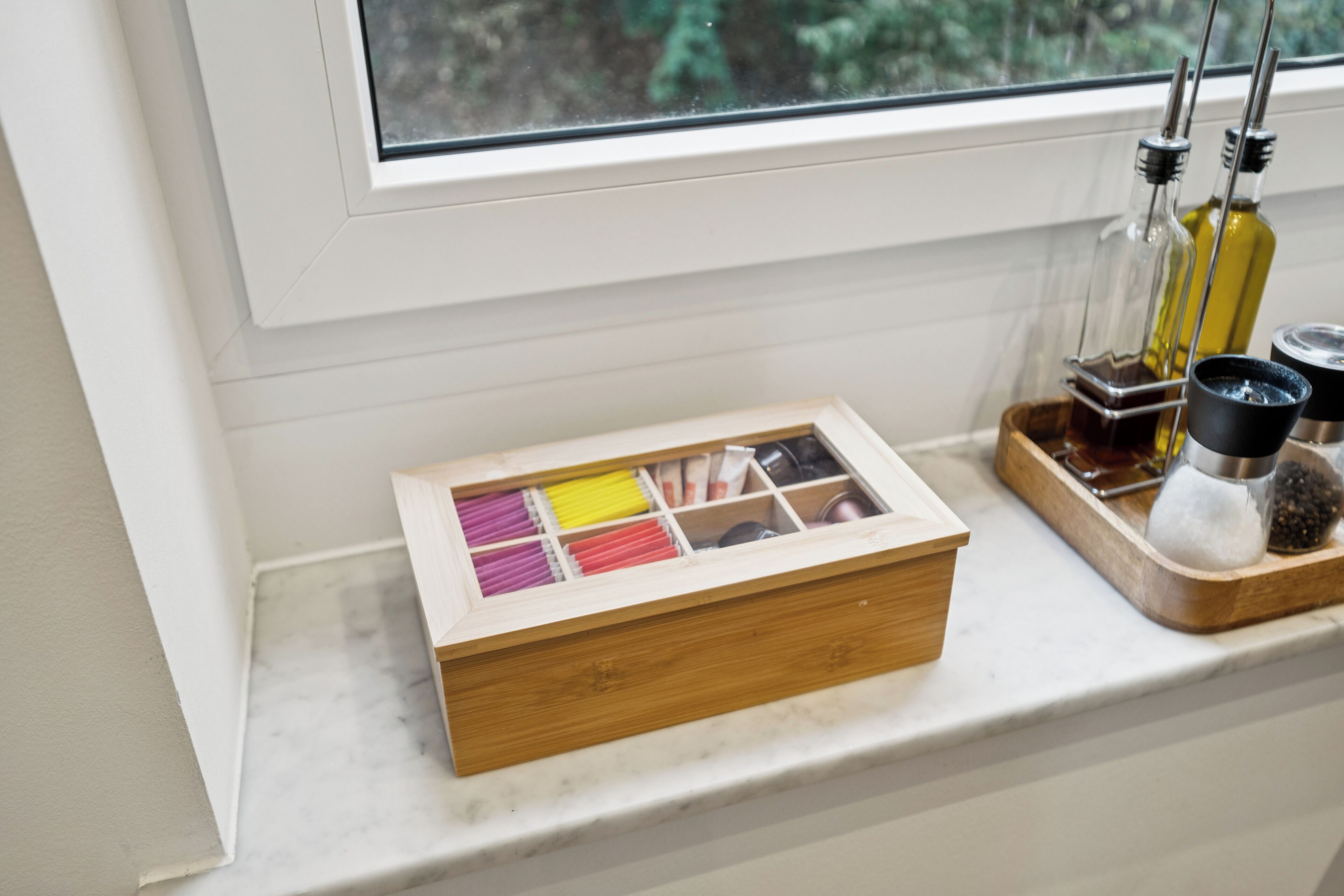 Une boîte en bois, divisée en compartiments, posée sur le rebord d’une fenêtre, à côté d’un plateau contenant des bouteilles et des condiments.