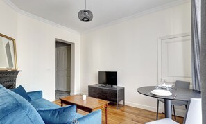 Ein modernes Wohnzimmer mit einem blauen Sofa, einem Holztisch, einer dunklen Kommode mit Flachbildfernseher und einem Esstisch für zwei Personen.
