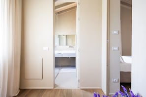 A room with a wooden floor, a bed, a bathroom with a mirror, and a door leading to another room.