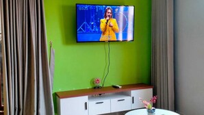 A modern living room with a green wall, a flat-screen TV mounted above a wooden cabinet, a round coffee table with a blue patterned rug, and a grey curtain.