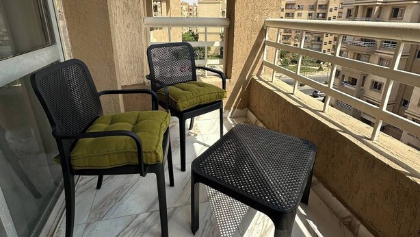 Ein Balkon mit zwei Stühlen, einem kleinen Tisch, einer Klimaanlage und Blick auf Wohnhäuser.