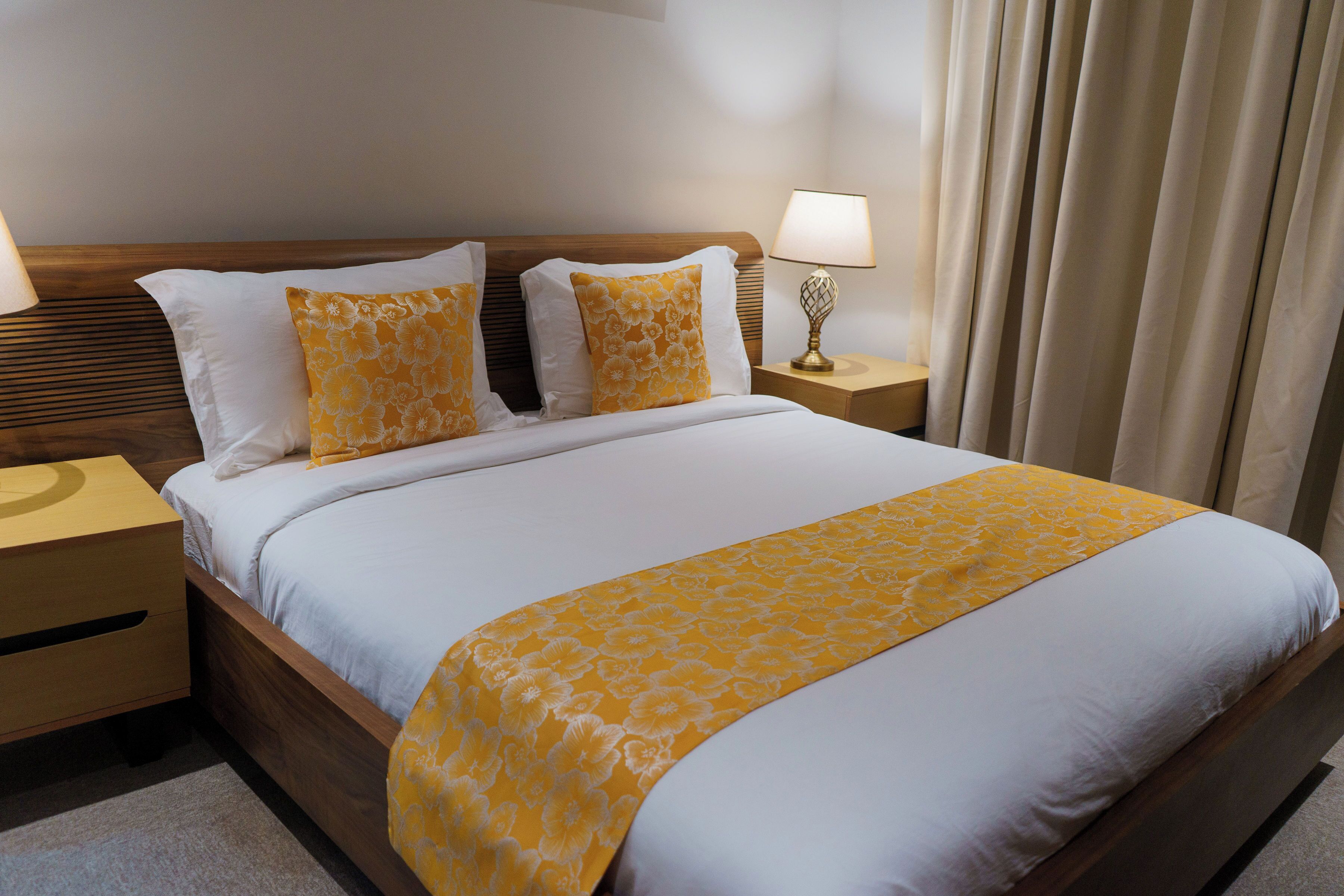 Een net opgemaakt bed met een wit en geel bloemenbedover, twee bijpassende kussens, een houten hoofdbord, een nachtkastje met een lamp en een raam met gordijnen.