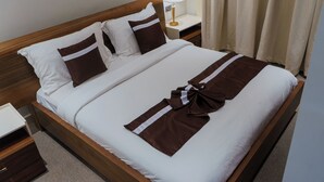 A neatly made bed with a brown and white bedspread, two pillows, a bedside table with a lamp, and a nightstand. - Staycation Villas (Dodoma)