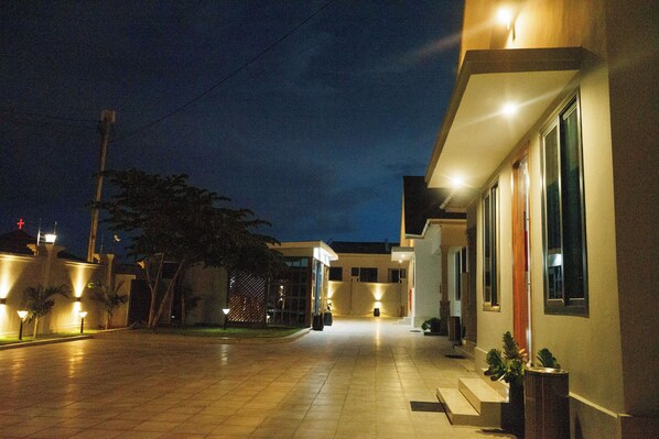 A nighttime view of a modern building with a well-lit entrance and a tiled pathway leading to the main entrance.