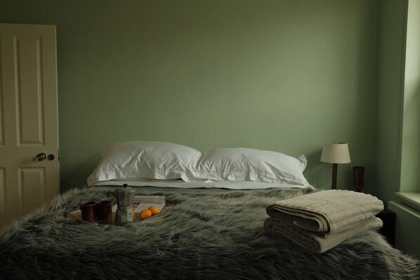 Un lit avec du linge de lit blanc, une couverture grise moelleuse, une serviette pliée et une table de chevet avec une lampe et un petit récipient.