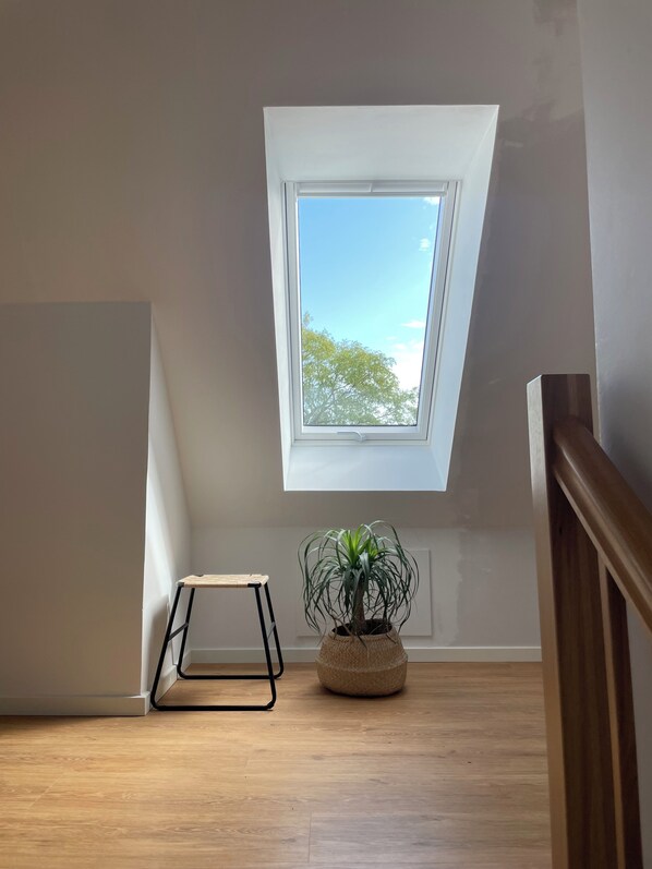 Una habitación con techo inclinado, tragaluz, planta en maceta y un taburete pequeño.