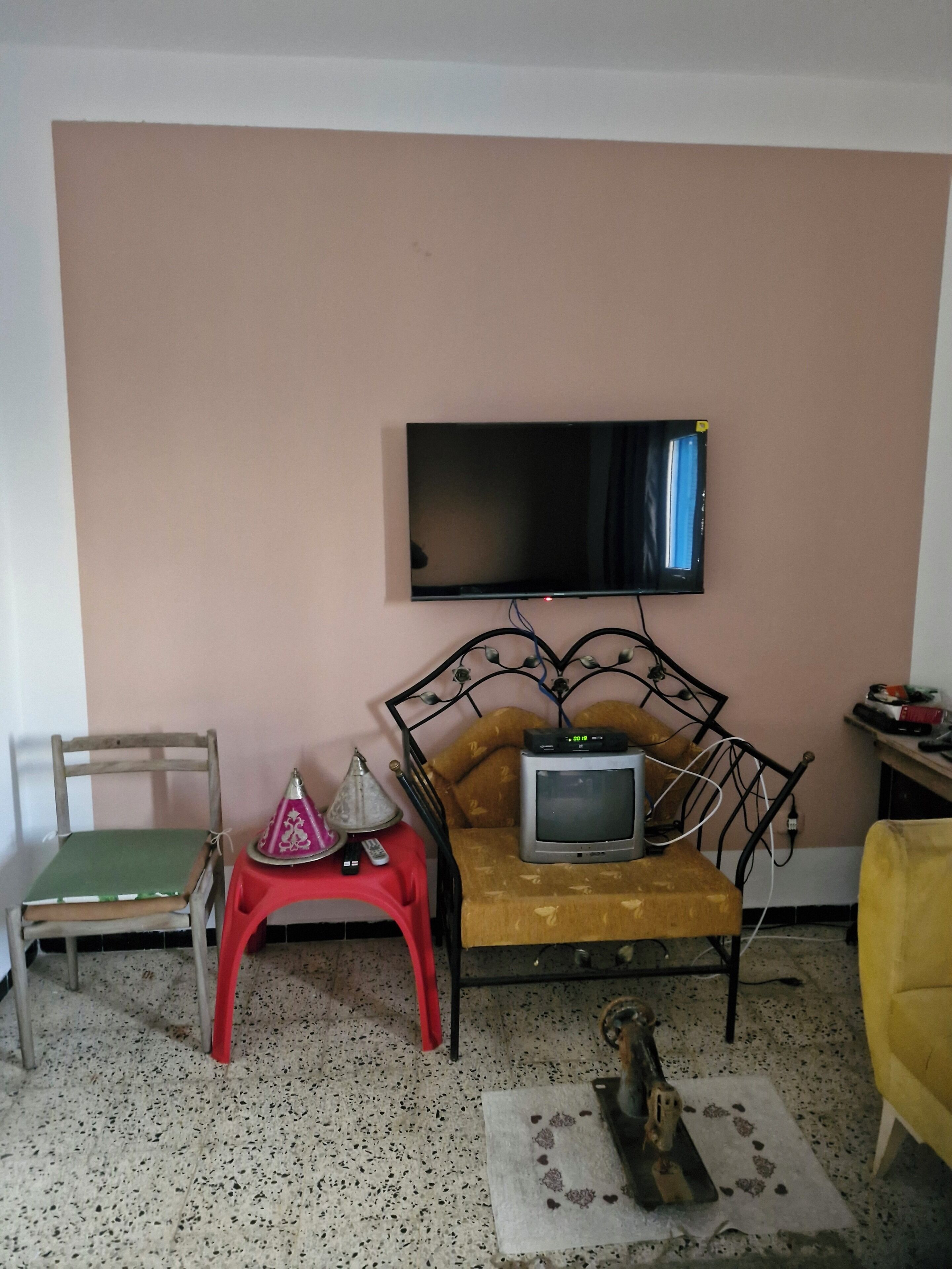 A room with a television mounted on the wall, a sofa with a small television, a wooden chair, a red stool, and a yellow armchair.