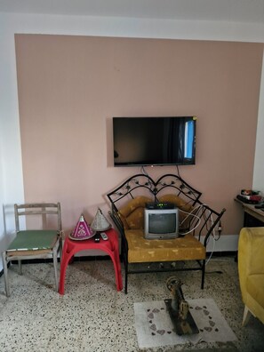 A room with a television mounted on the wall, a sofa with a small television, a wooden chair, a red stool, and a yellow armchair. - Spacious, pleasant blue apartment with large balcom, tunis ibn cina (Tunis)