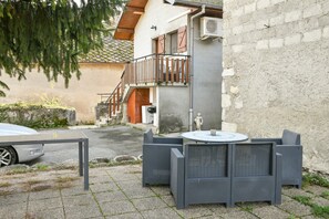 Un patio con mobiliario de mimbre, un coche estacionado cerca y un edificio con balcón y escalones.