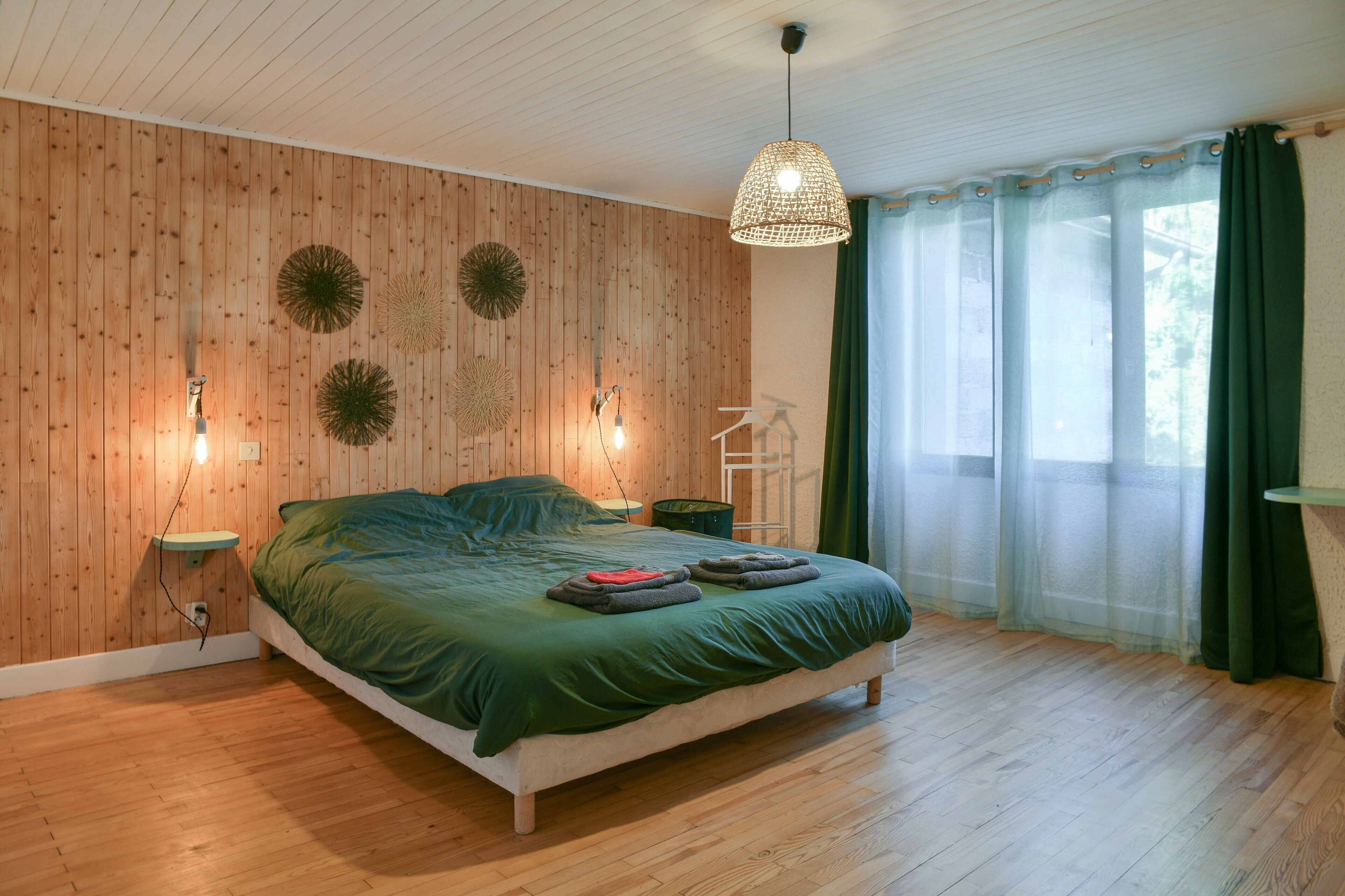 A bedroom with a wooden wall, a bed with green bedding, a hanging light fixture, and a window with sheer curtains.