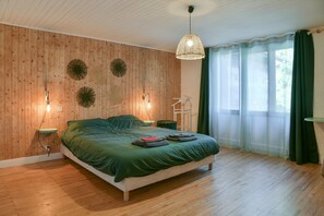 Un dormitorio con pared de madera, una cama con ropa de cama verde, una lámpara colgante y una ventana con cortinas traslúcidas.