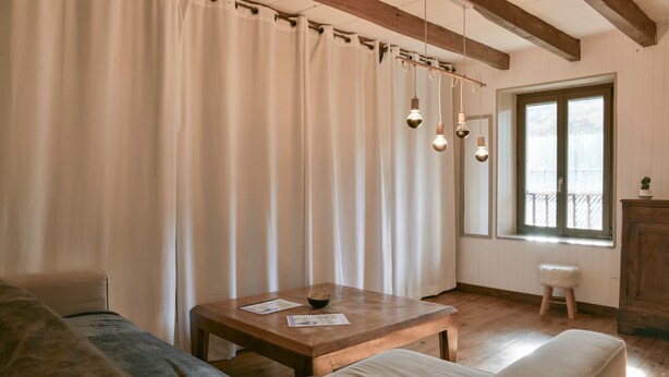 Une pièce avec une table en bois, un canapé, un petit tabouret et une fenêtre avec des rideaux.