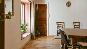 Une pièce avec une table à manger en bois, deux chaises, une porte en bois, une fenêtre avec une plante et un miroir au mur.