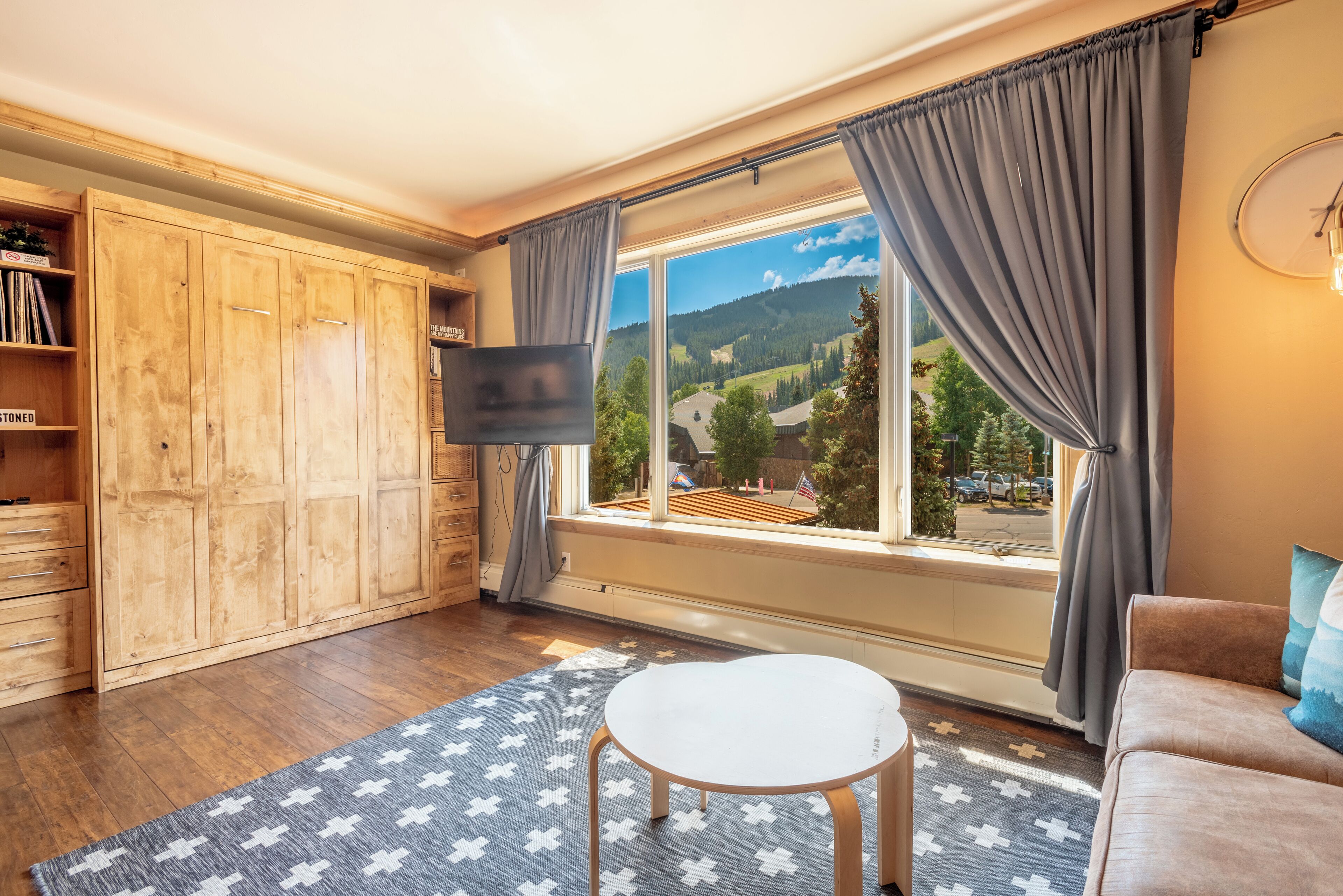 A room with a large window, wooden wardrobe, a flat-screen TV, a round table, and a sofa.