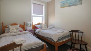 A bedroom with two wooden beds, a nightstand, a chair, and a window with blinds. - Pebbles (Eastbourne)