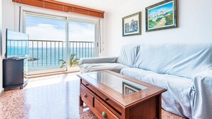Una sala de estar con un sofá, una mesa de centro y un televisor. Se ve una ventana grande con vistas al mar.