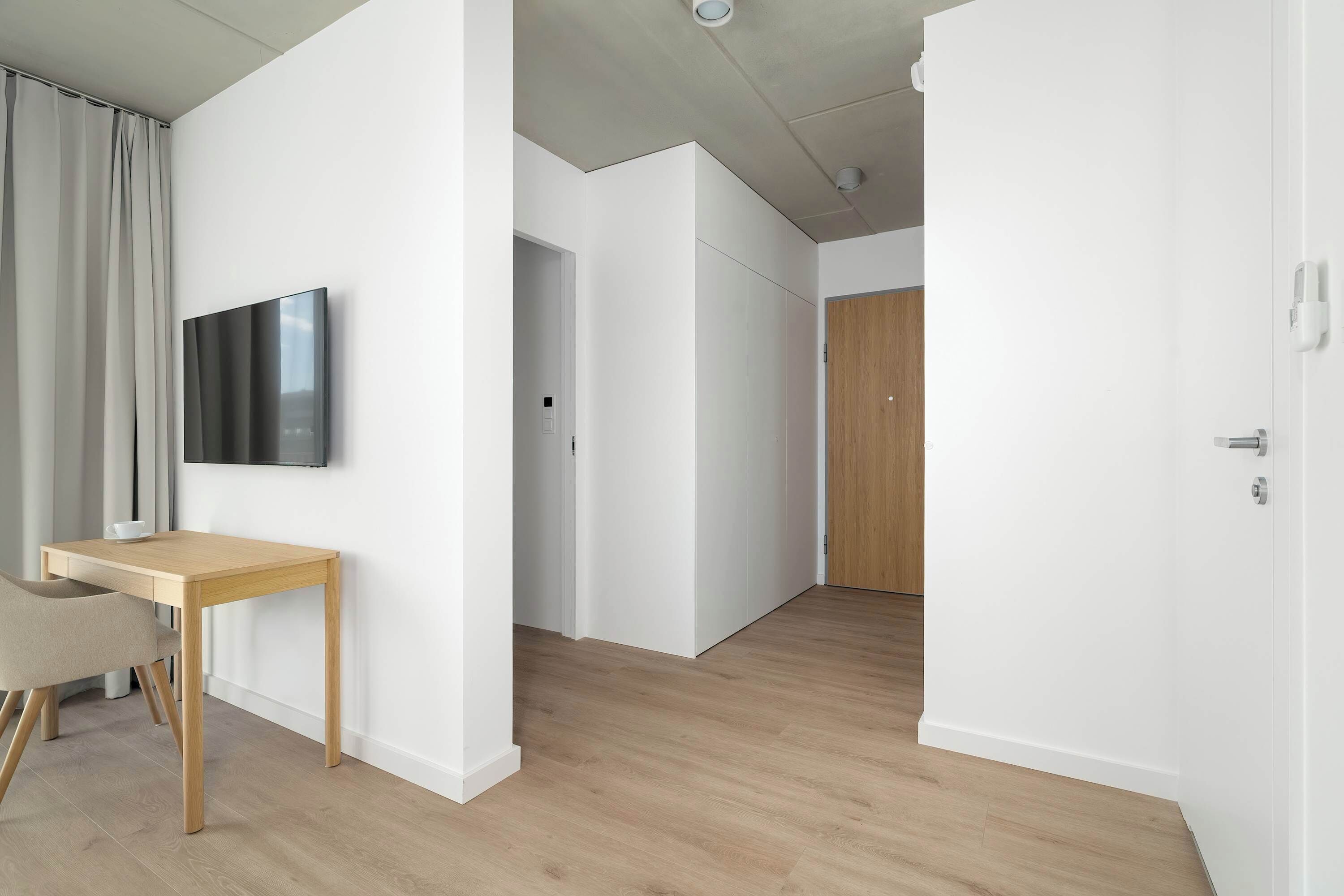 Ein modernes, minimalistisches Zimmer mit Holzboden, einem an der Wand montierten Flachbildfernseher, einem kleinen Holztisch und einer Tür zu einem anderen Raum.