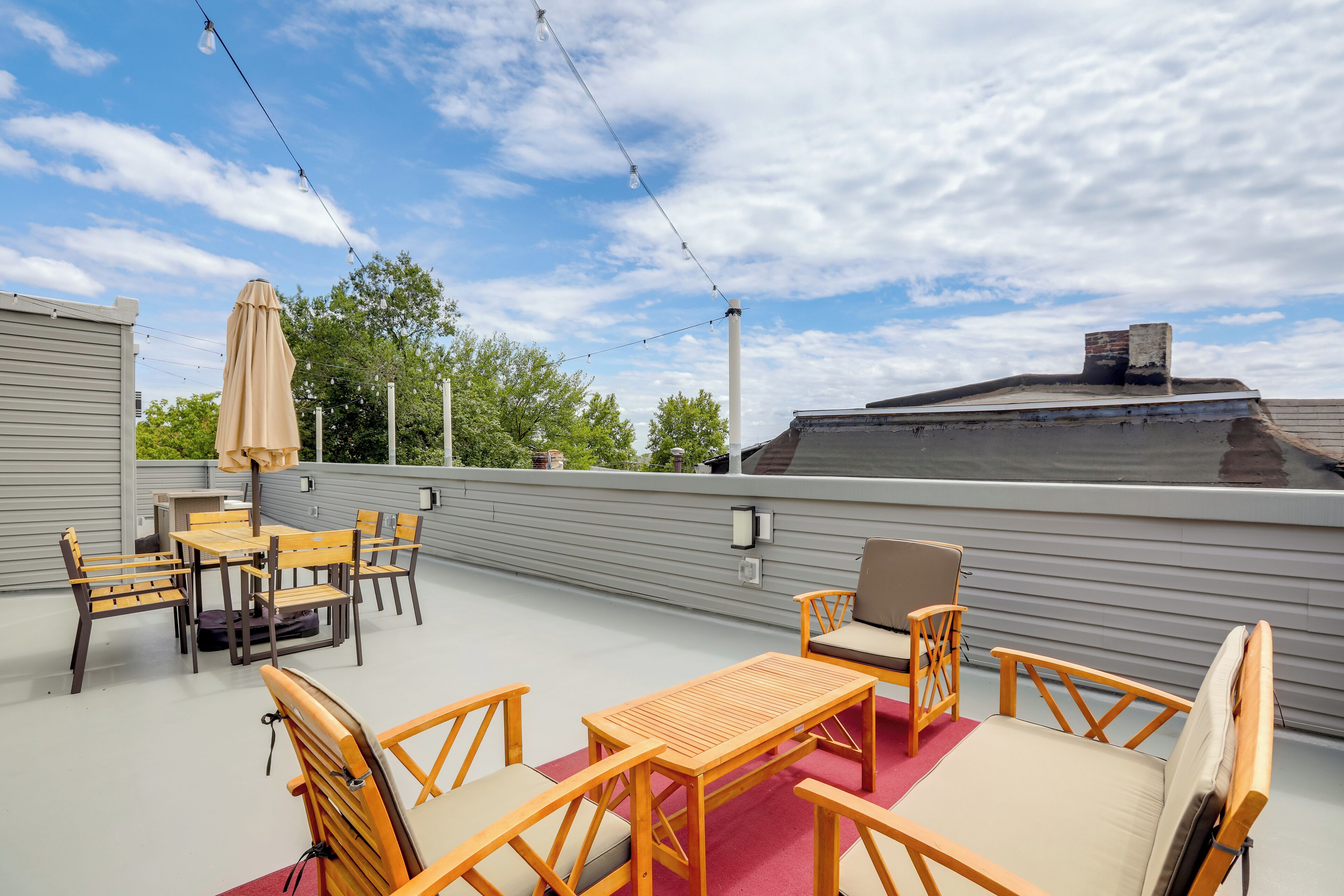 A rooftop patio with wooden furniture, string lights, and a view of the sky and trees.