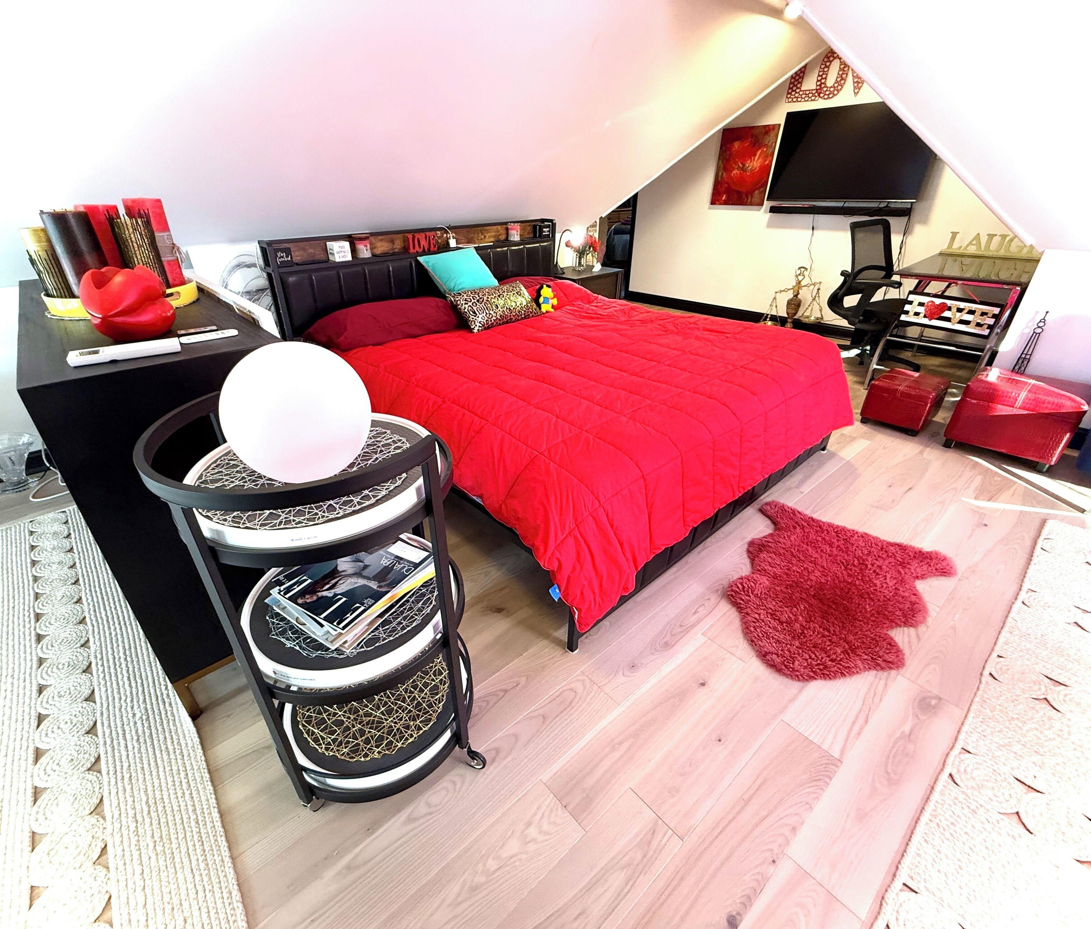A bedroom with a red bedspread, a black headboard, and a small round table with a white lamp.