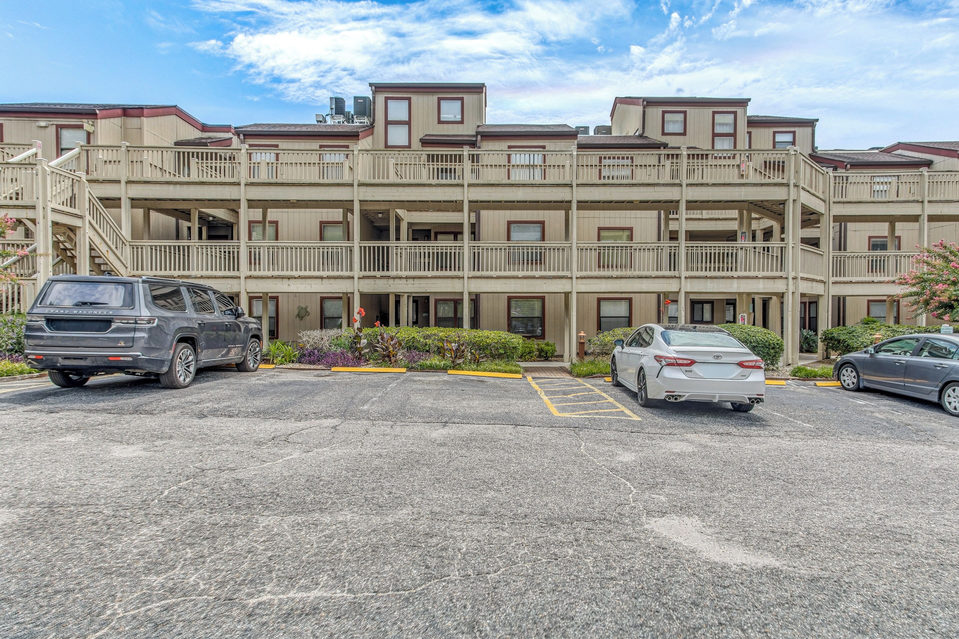 A multi-story parking garage with a row of parked cars, a stairway to the upper levels, and a building with balconies in the background.