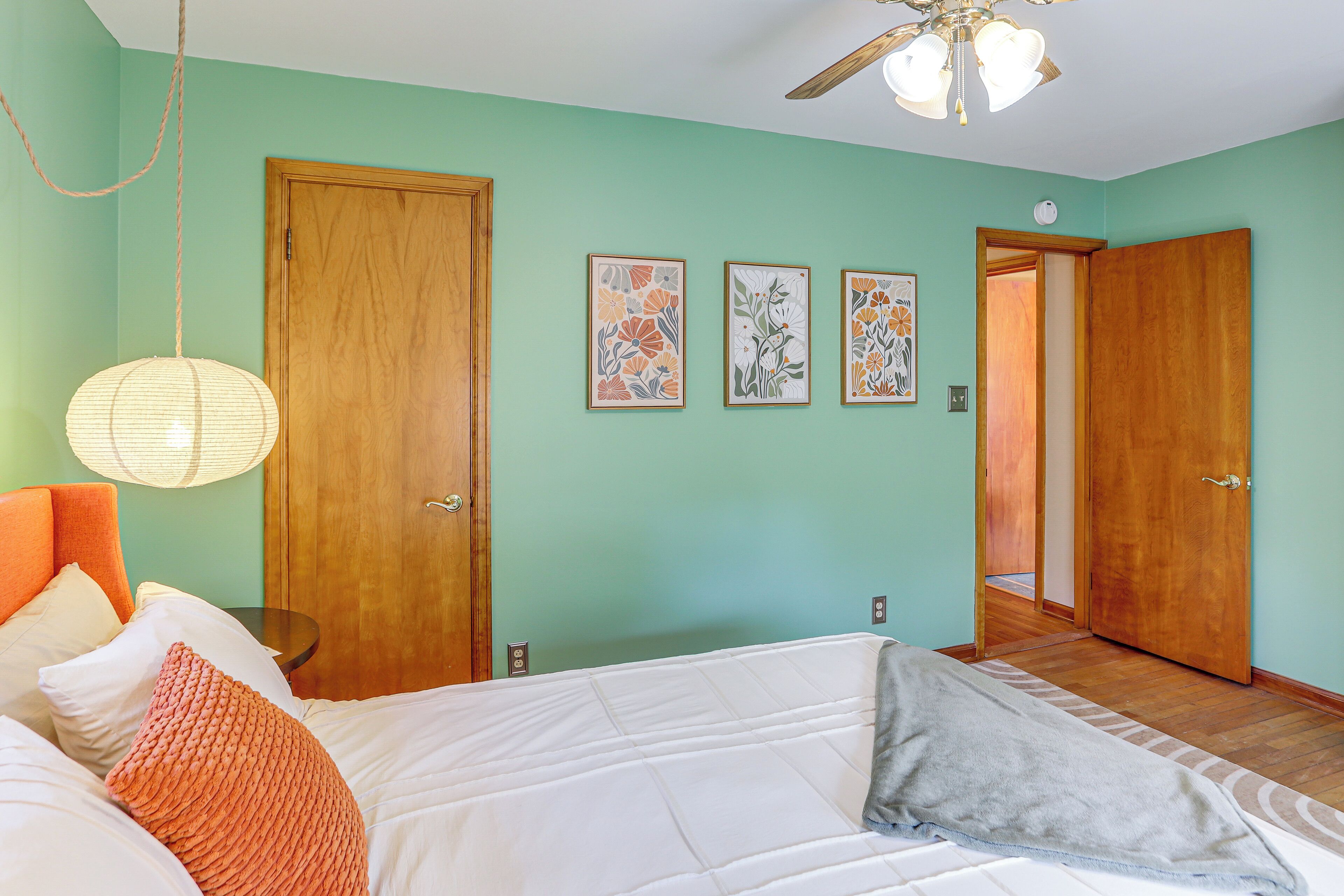 A bedroom with a bed, pillows, a hanging lamp, and three framed artworks on the wall.