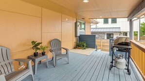A covered outdoor area with a wooden deck, two Adirondack chairs, a grill, and potted plants.