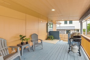 Uma área externa coberta com deck de madeira, duas cadeiras Adirondack, churrasqueira e vasos com plantas.