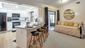 A modern kitchen with white cabinets, a central island with stools, a microwave, and a coffee machine. There is a sofa with a quilted cover and a large abstract painting on the wall. - Fishing Dock & Fire Pit! Gulf Cove Group Getaway (Port Charlotte)
