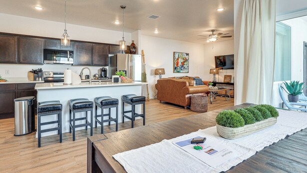A modern kitchen with dark wood cabinets, a central island with stools, a stainless steel trash can, and a dining area with a wooden table and a decorative plant. - Home w/ Private Patio 10 Mi to Rio Grande! (Las Cruces)