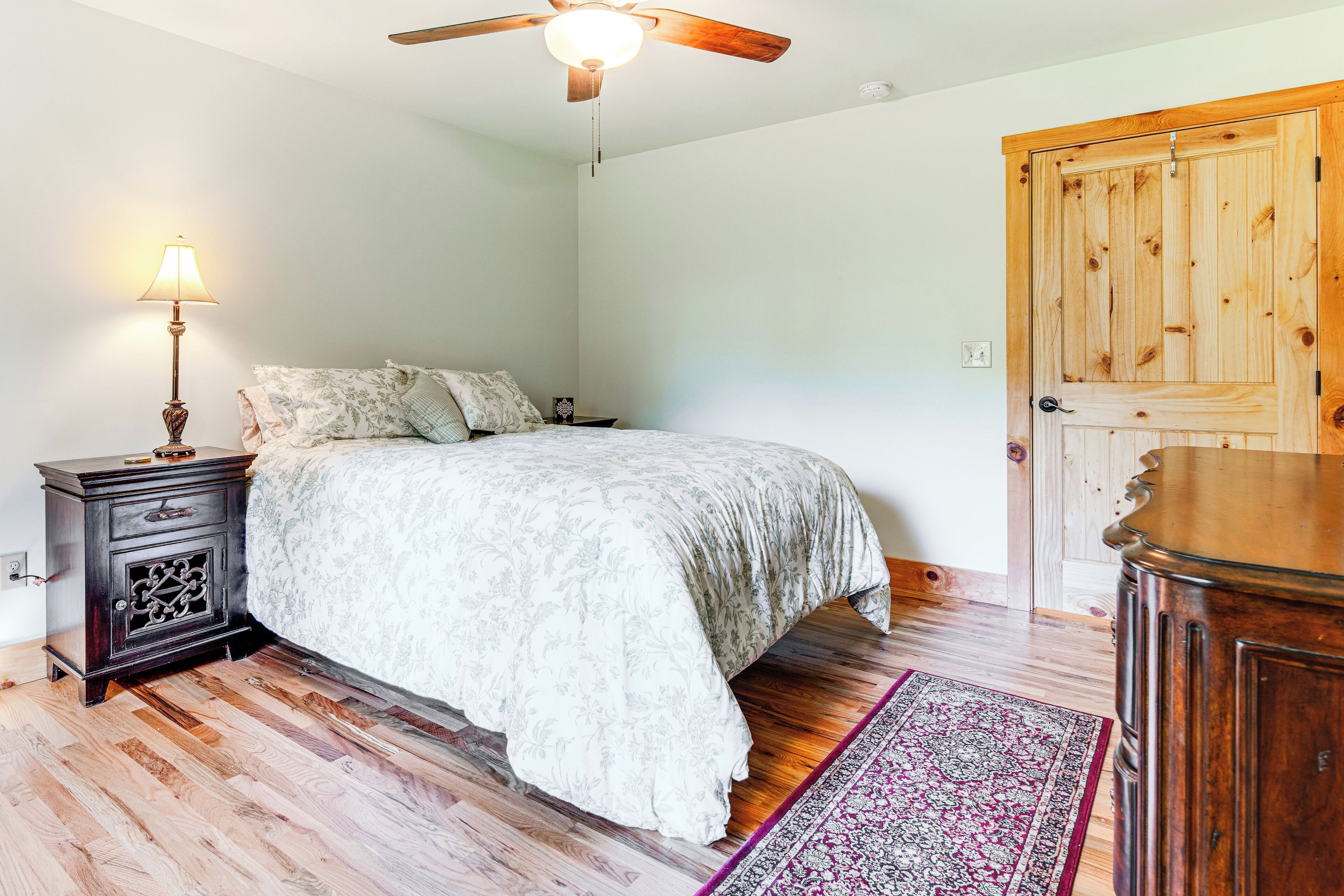 A bedroom with a bed, wooden floor, a nightstand, a lamp, a ceiling fan, and a door.