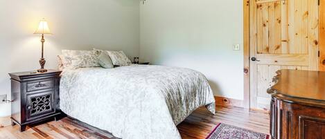 A bedroom with a bed, wooden floor, a nightstand, a lamp, a ceiling fan, and a door.