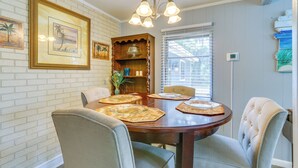 A dining room with a brick wall, a wooden dining table, four upholstered chairs, a chandelier, and framed artwork.
