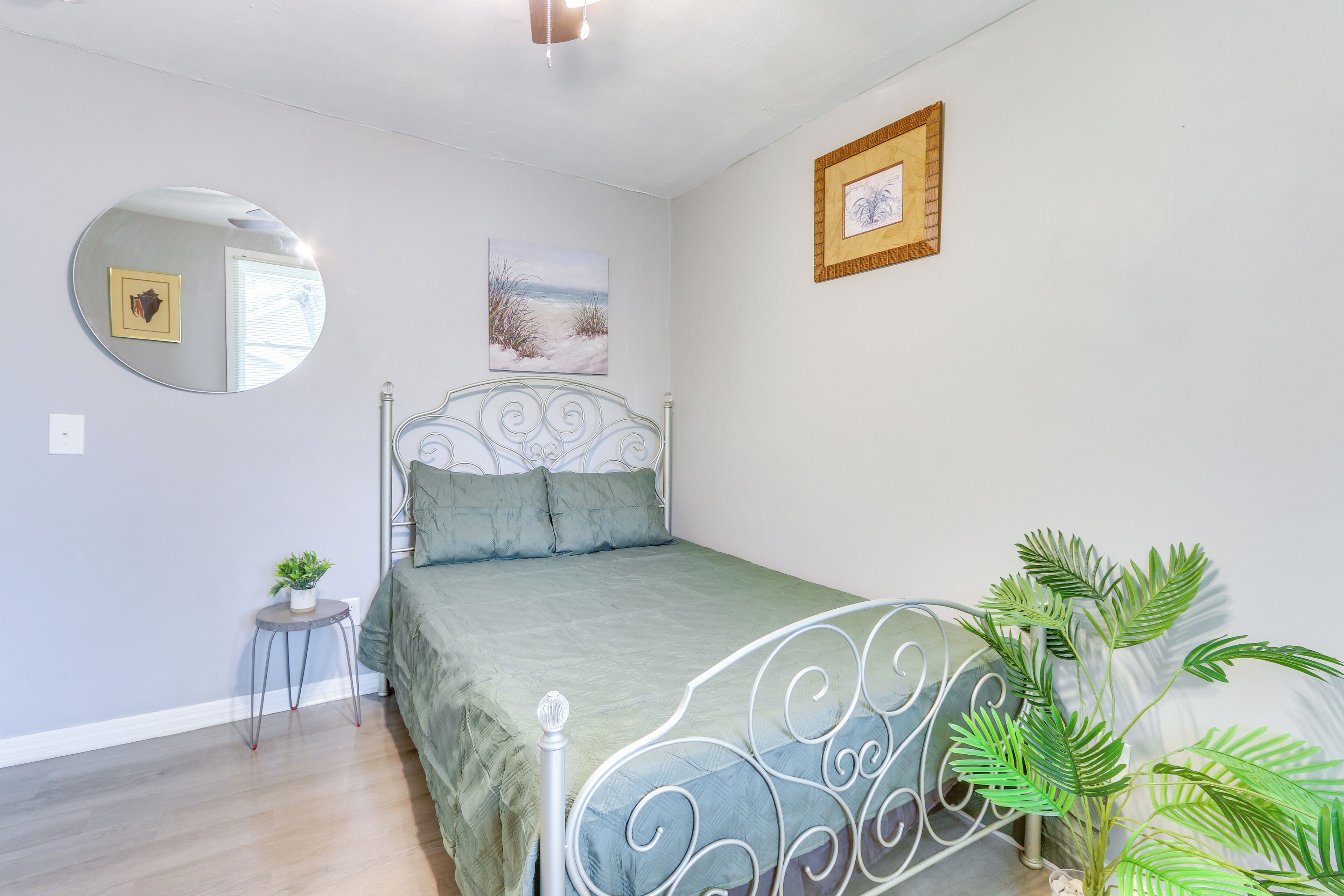 Quarto com cama, espelho redondo, pequena mesa com planta e quadro na parede.
