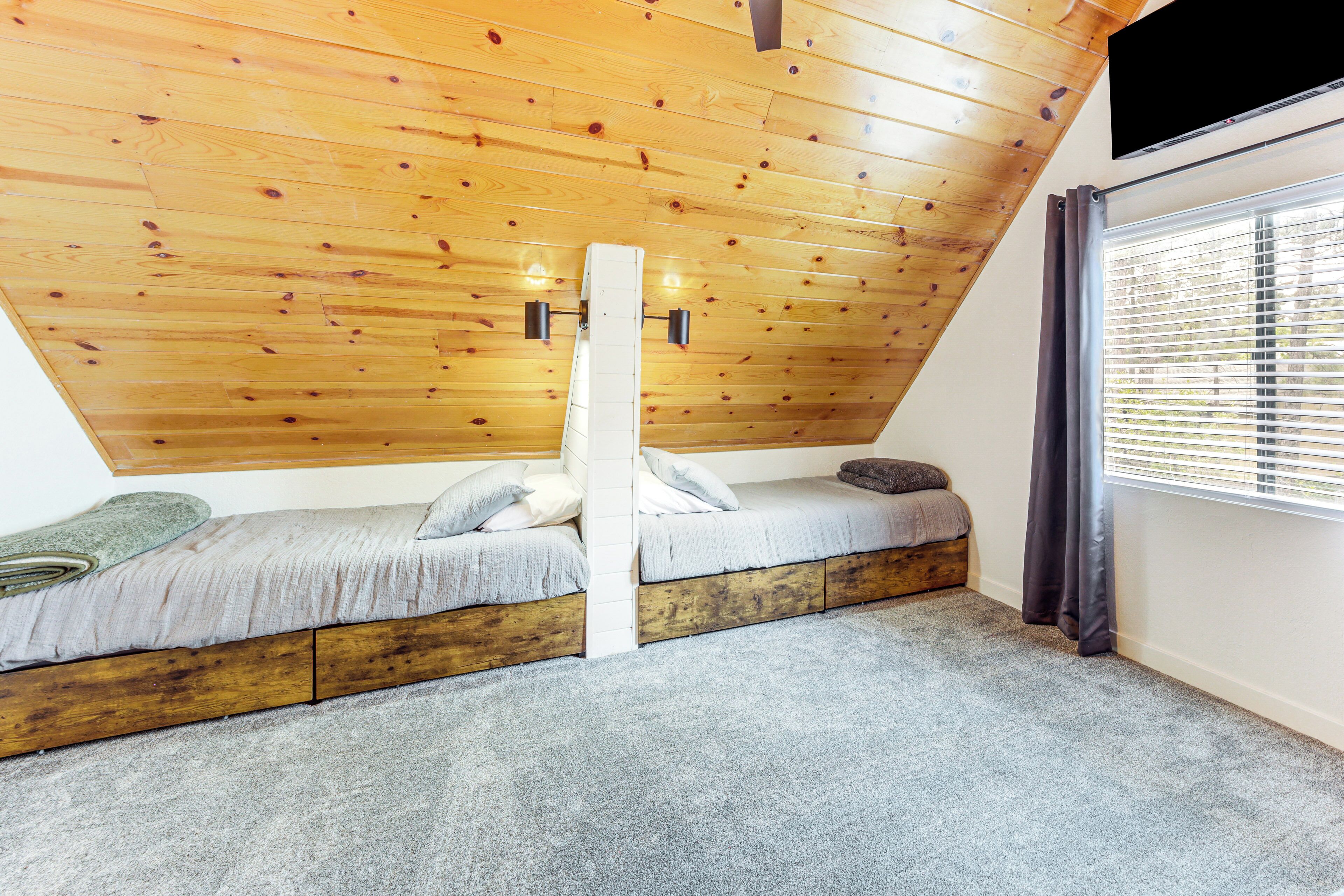 Une chambre située sous les toits, avec un plafond en pente, deux lits, une grande fenêtre avec stores et un téléviseur fixé au mur.
