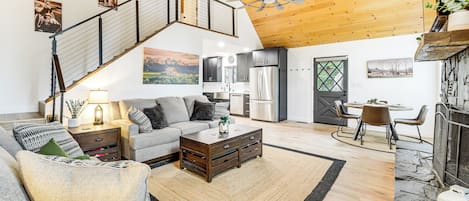 A spacious living area with a high ceiling, wooden beams, a staircase, a fireplace, and a dining area with a view of the kitchen.