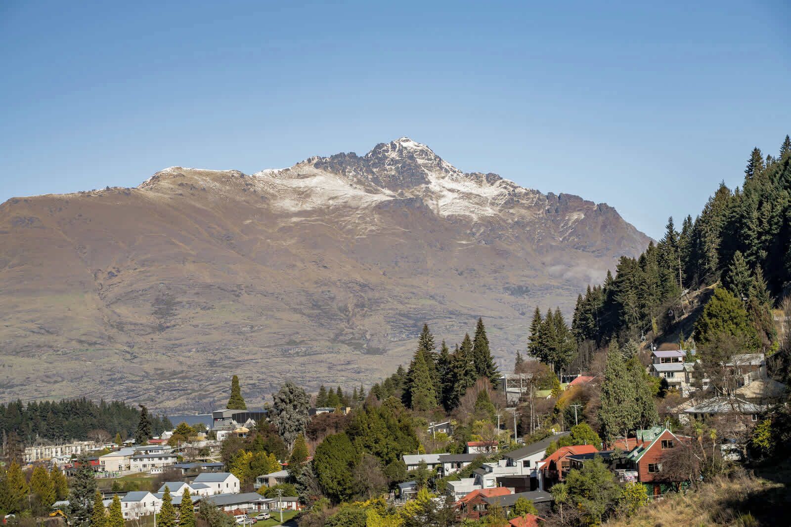 A mountain with snow-capped peaks in the background, surrounded by a residential area with houses and trees.