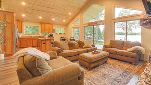 A spacious living area with a large sectional sofa, a coffee table, and a fireplace. There is a kitchen in the background with wooden cabinets and a large window. - Boat Dock, BBQ & Fireside Fun: Clam Lake Cabin (Siren)